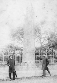 General view of face of Sueno's Stone. A bearded and a moustachioed gentleman stand in front of the elaborate cast-iron railings.
Photographic copy of cabinet print of Sueno's Stone: the mount bears the printed inscription 'With the Seasons Greetings'.