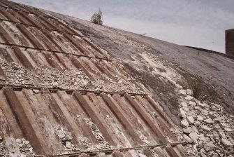 Detailed view from SE of centre part of hangar, showing exposed fabricated steel shell (left), with adjacent relatively intact outer layer of concrete, covered with bitumen