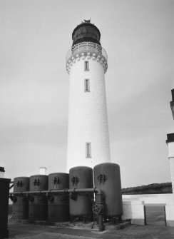 Detailed view from SE of lighthouse tower, with battery of five dome-ended air receivers (in the foreground) which stored compressed air (generated in the fog-horn engine house to left) to power the fog horn.  Note the two receivers to the right are welded, and the three to the left are riveted.