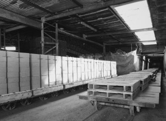 Interior view from NW of bogie carrying cement bricks in foreground, with storage shed (previously an aircraft hanger re-erected at the site) in background
