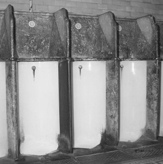 Interior. Detail of three of the 23 stall urinals, showing the white-glazed fireclay stalls with simulated grey marble winged hoods.  These examples retain their Doulton (of Lambeth, London, and Paisley) nameplates