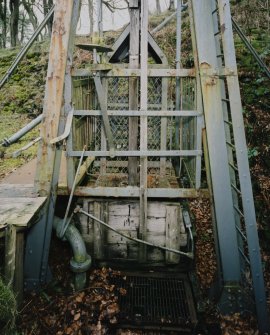 Detailed view from SW of the top of the shaft, showing the cage, and in the foreground (covered with a grille) the escape route, which comprises twelve ladders running parallel to the shaft.  The mechanism attached to the left upright of the headframe is the bell allowing the winding-engine operator to communicate with the men at the pit bottom.