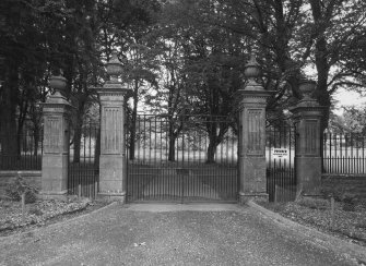 View from S showing set of four Edwardian classical stone gatepiers topped with urns with gates and flanking railings.