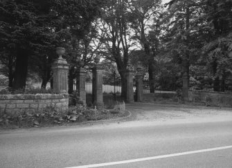 View from SW showing set of four Edwardian classical stone gatepiers topped with urns with gates and flanking railings.