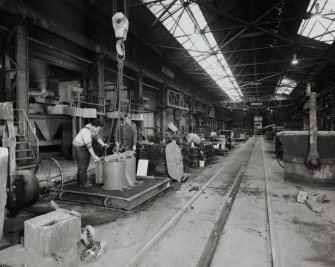 General interior view from SE down main moulding bay of foundry, with furnaces (left) and mould being prepared in foreground, and rail-mounted sand-filling machine in distance