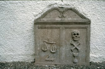 View of headstone  to AF  IF with scales, skull, crossbones against wall of Glendevon Parish Church.