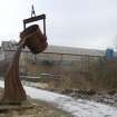 Voew of sculpture alongside public path of line of dismantled railway with back view of foundry buildings from south.