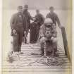 Diver in diver suit sitting on the pier with a group of men at Dalerb Slip, Kenmore', Loch Tay. 1890. Equipment and ropes to guide the diver can be seen.

