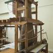 Interior. General view. Jaquard loom in gallery area. The warp threads in tension on the loom frame are clearly visible as is the jacquard device above the loom which manages the weft threads (by means of punched cards) to create the structure and pattern of the cloth being woven. The holes and blanks on the cards allow the hooks in the frame to select or not select threads to create the complex patterns in the cloth