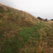 View from the ENE of the NNW scarp of the blast mound surrounding the central Final Wash House (Final Wash House W30). The entrance tunnel for the narrow gauge railway lies just behind the post, while the gash defined by darker vegetation at the foot of the scarp (foreground) marks the site of a safety shelter