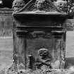 View of gravestone of David Sevitch.
Skulls on top slopes, winged cherub in pediment, tailor's 'goose' and shears.