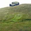 Oblique aerial view centred on the remains of the roundhouse, buildings, field banks and lazy beds, with the settlement mound adjacent and a peat stack stand in the foreground, Skipisdale, Mingulay, taken from the NE.