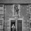 South elevation of central portion of Alexander Selkirk's former house, with woman standing in doorway and 1885 statue of the mariner from Fife above.