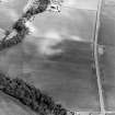 Oblique aerial view centred on cropmarks of an enclosure, with a Roman road adjacent, taken from the NW.