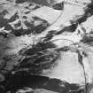 Oblique aerial view centred on the remains of buildings and enclosures with road bridge, rig and farmsteading adjacent, taken from the NW.