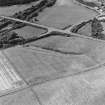 Oblique aerial photograph of Glenluce taken from the SE, centred on the cropmarks of a Roman temporary camp.