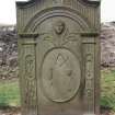 Forgan, St. Fillans Churchyard.
General view of gravestone with arched top with central rosette. Very finely carved with fluted pilasters, each with rosettes and roses inset. Arched pediment with head and thistles below. Oval panel with dividers, set-square, pick, mallet, trowel and hammer.