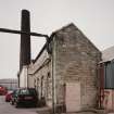 View from SE of SE end of boilerhouse, also showing the associated chimney stack in the background.  Part of the boilerhouse wall, centre, beneath chimney, has had to be replaced with a crimped steel shell.