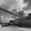 View from W showing overhead conveyor from primary crusher (base visible to right) to former shaft kilns (now storage hoppers to left).  The supporting strut for the conveyor is a surviving portion of one of the original conveyors supplying the shaft kilns