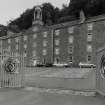 View from S of New Buildings, with cobbled area in the foreground delineating the position of the former Mill Offices, which were demolished in the 1980s.  Also in the foreground are the main gates to the mills, which bear plaques commemorating the recent World-Heritage Inscription by UNESCO