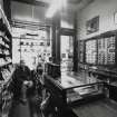 View of interior of tobacconists shop from N (behind counter) showing shop entrance and window, display cases containing pipes and shop assistant seated behind counter